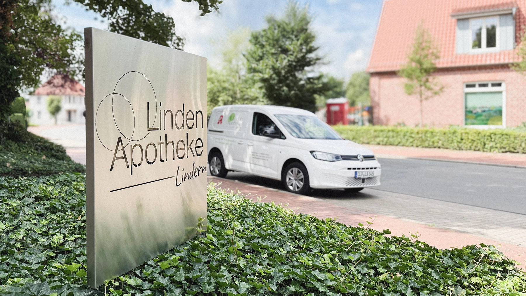 Vor einem Gebäude mit rotem Ziegeldach steht ein weißer Lieferwagen mit Apotheken-Logo, im Vordergrund ein Schild mit der Aufschrift „Linden Apotheke Lindern“, umgeben von grünem Efeu.