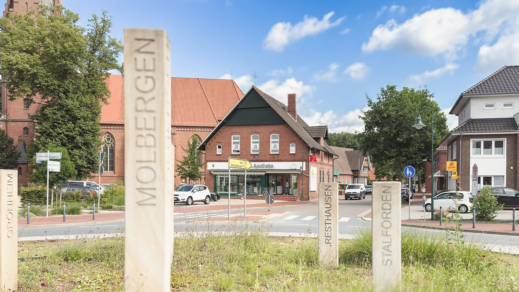 Straßenansicht der Johannes Apotheke in Molbergen an einer Kreuzung, umgeben von Bäumen und Gehwegen, bei sonnigem Wetter.