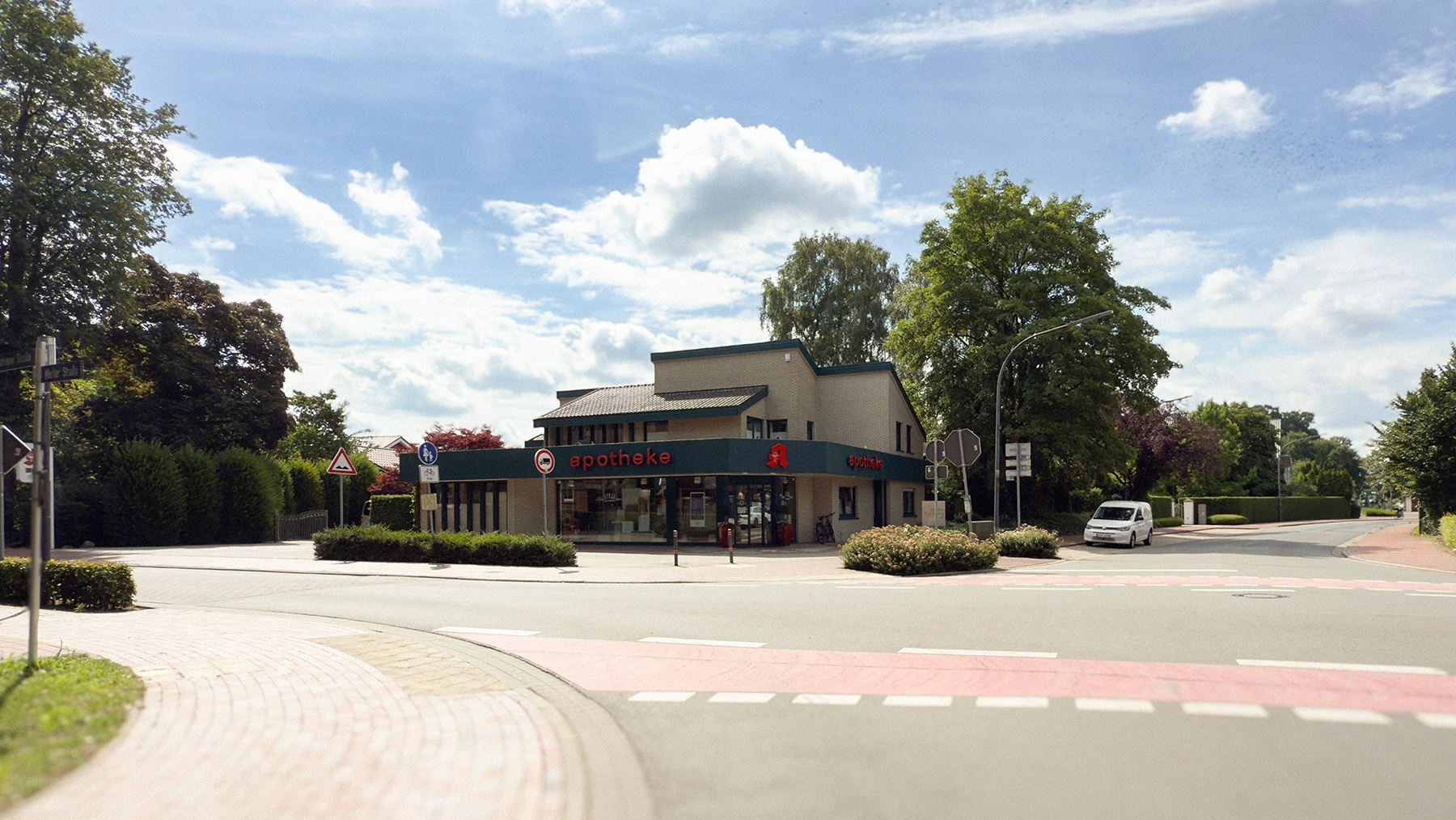 Straßenansicht der Linden Apotheke in Lindern an einer Kreuzung, umgeben von Bäumen und Gehwegen, bei sonnigem Wetter.